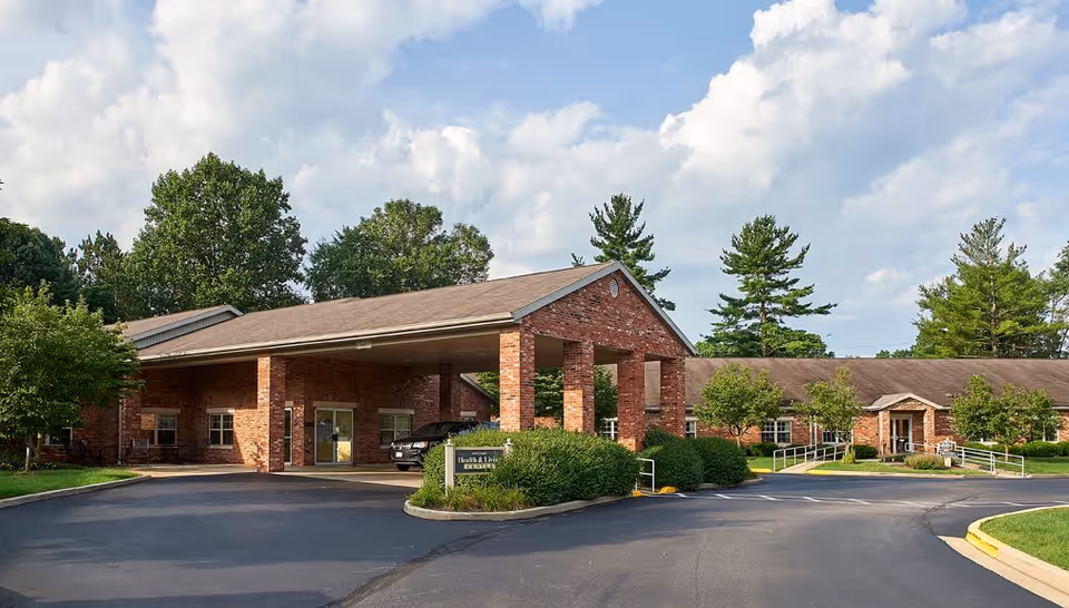 Exterior view of a single-story brick senior living facility building with a covered entrance driveway, surrounded by trees and bushes under a partly cloudy sky.