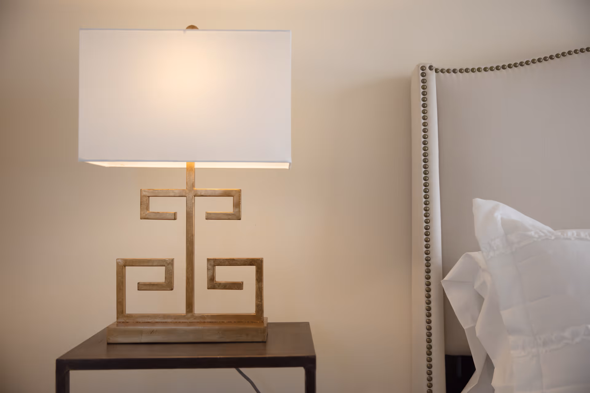 A close-up view of a bedside table with a decorative lamp featuring a geometric gold base and a rectangular white lampshade. To the right, part of a beige upholstered headboard with nailhead trim and white pillows is visible.
