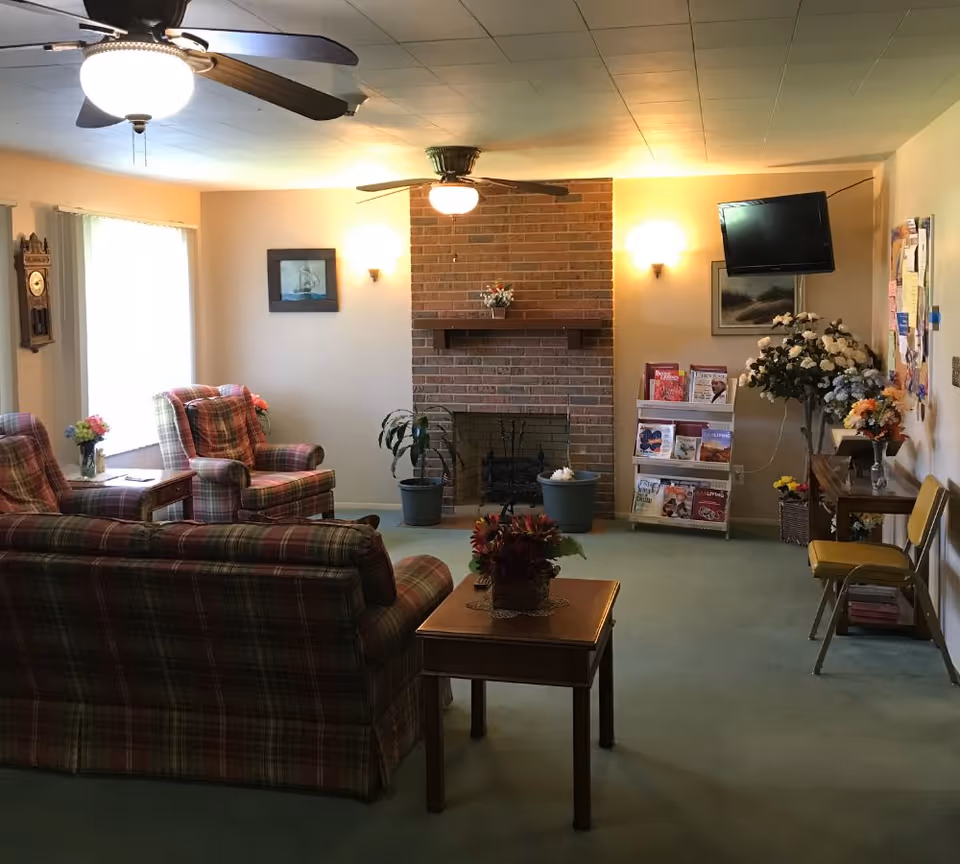 A cozy senior living common area with plaid upholstered chairs and sofa, a wooden coffee table with a flower arrangement, a brick fireplace with potted plants on either side, a wall-mounted TV, a magazine rack, and floral decorations. The room is softly lit with ceiling fans and wall sconces.
