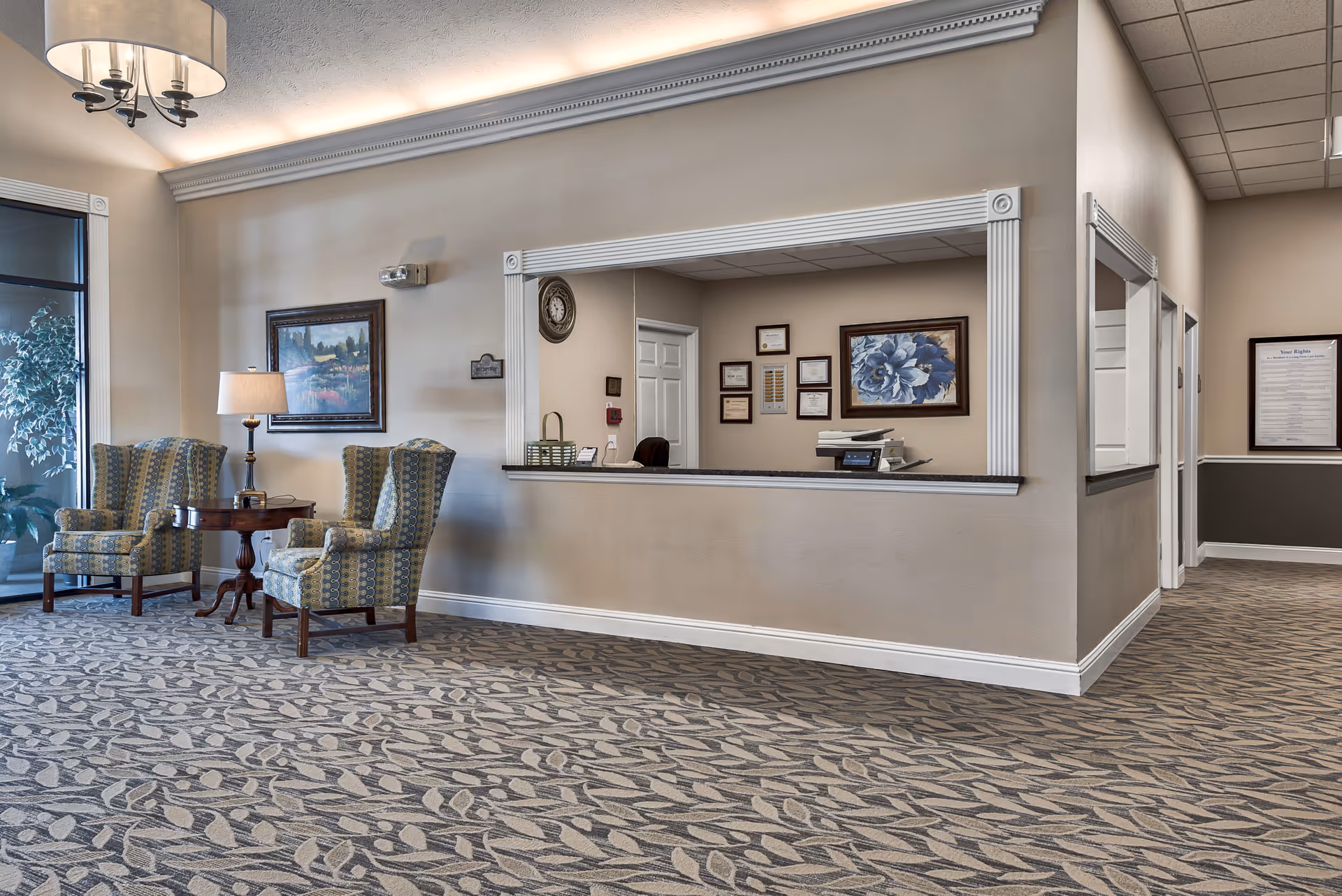 A senior living reception lobby with two patterned armchairs and a small table beside a service window and framed artwork.