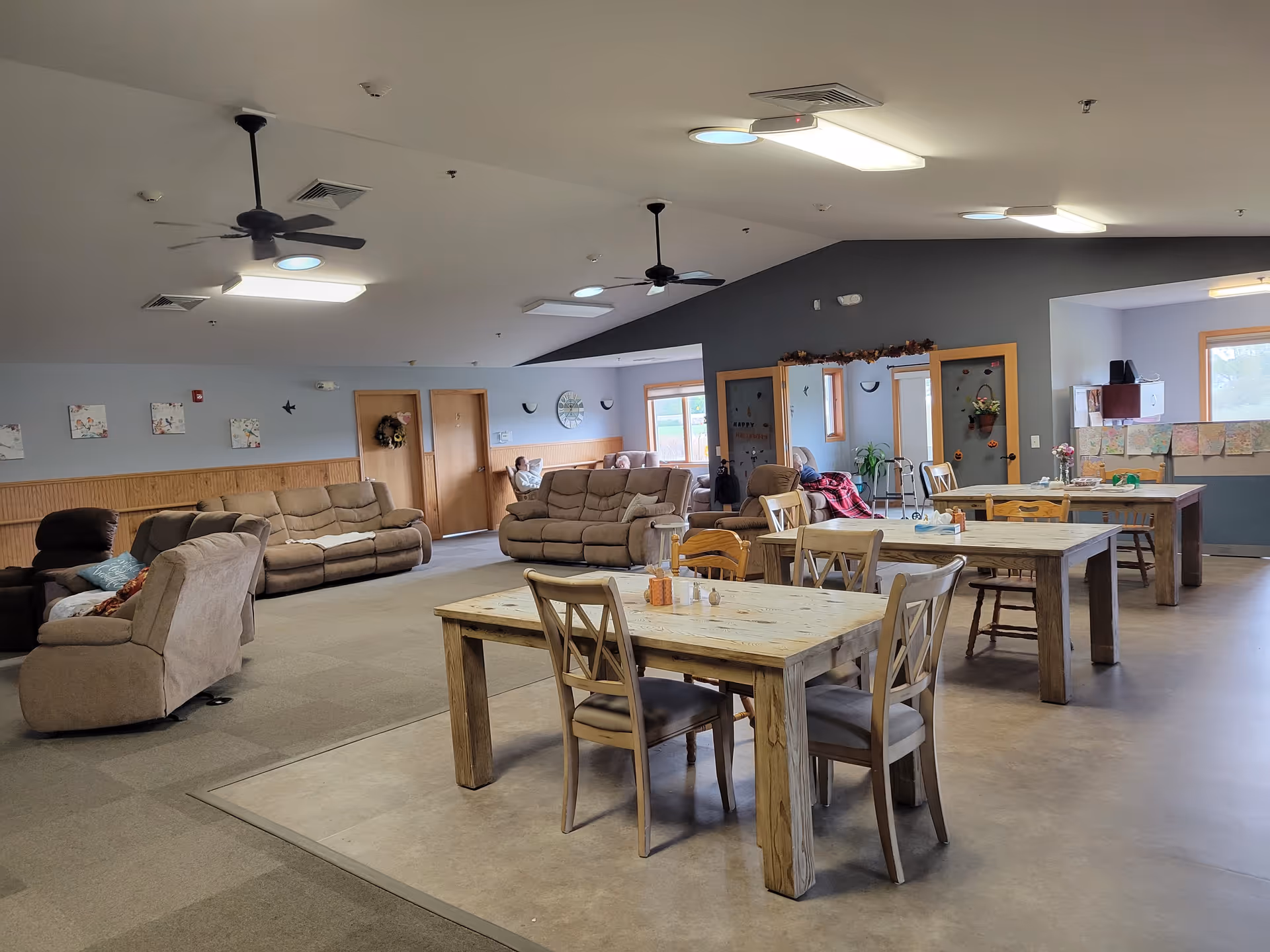 A spacious common area in Albany Oaks Assisted Living featuring several wooden tables with chairs in the foreground and multiple beige recliner sofas arranged along the walls. The room has ceiling fans, overhead lights, and windows letting in natural light. Decorations and artwork are visible on the walls, and a few residents are seated in the background.