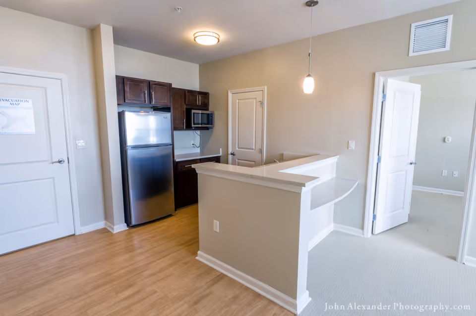 Interior view of a small kitchen area with wooden cabinets, a stainless steel refrigerator, a microwave, and a countertop with a breakfast bar. The room has light-colored walls and flooring, with an open doorway leading to another room with carpeted flooring.