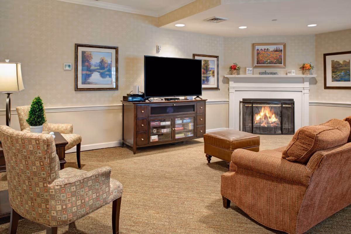 Cozy living room with a lit fireplace, a large flat-screen TV on a wooden cabinet, two patterned armchairs, a brown sofa, and a matching ottoman. The walls are decorated with framed landscape paintings and floral arrangements on the mantel.