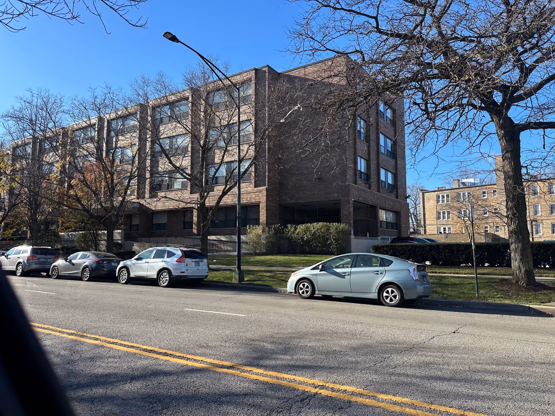 Exterior view of a multi-story brick building with several windows, situated along a street with parked cars and leafless trees under a clear blue sky.