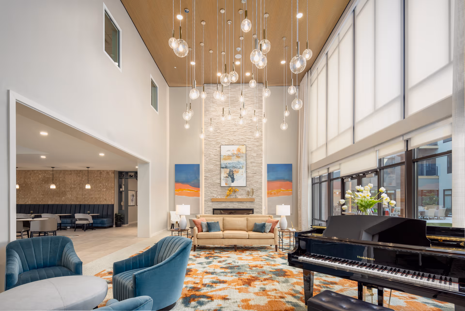 A spacious and modern living room area with high ceilings and large windows letting in natural light. The room features a beige sofa with colorful cushions, two blue armchairs, a round coffee table, a patterned area rug, and a black grand piano with a vase of white flowers on top. Pendant lights hang from the wooden ceiling, and there are two abstract paintings on the wall behind the sofa.