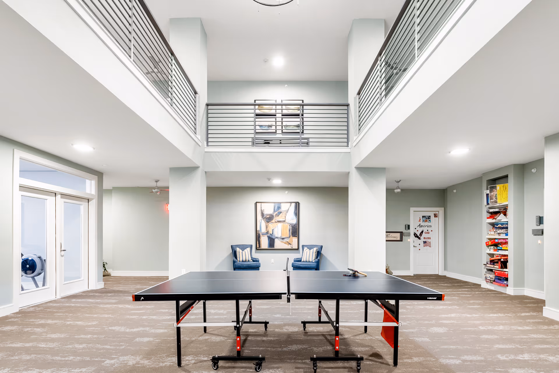 Interior view of a spacious recreational room with a ping pong table in the center, two blue armchairs against the back wall, a large abstract painting above the chairs, and shelves filled with board games on the right side. The room features a high ceiling with a balcony railing on the upper level and large glass doors on the left.