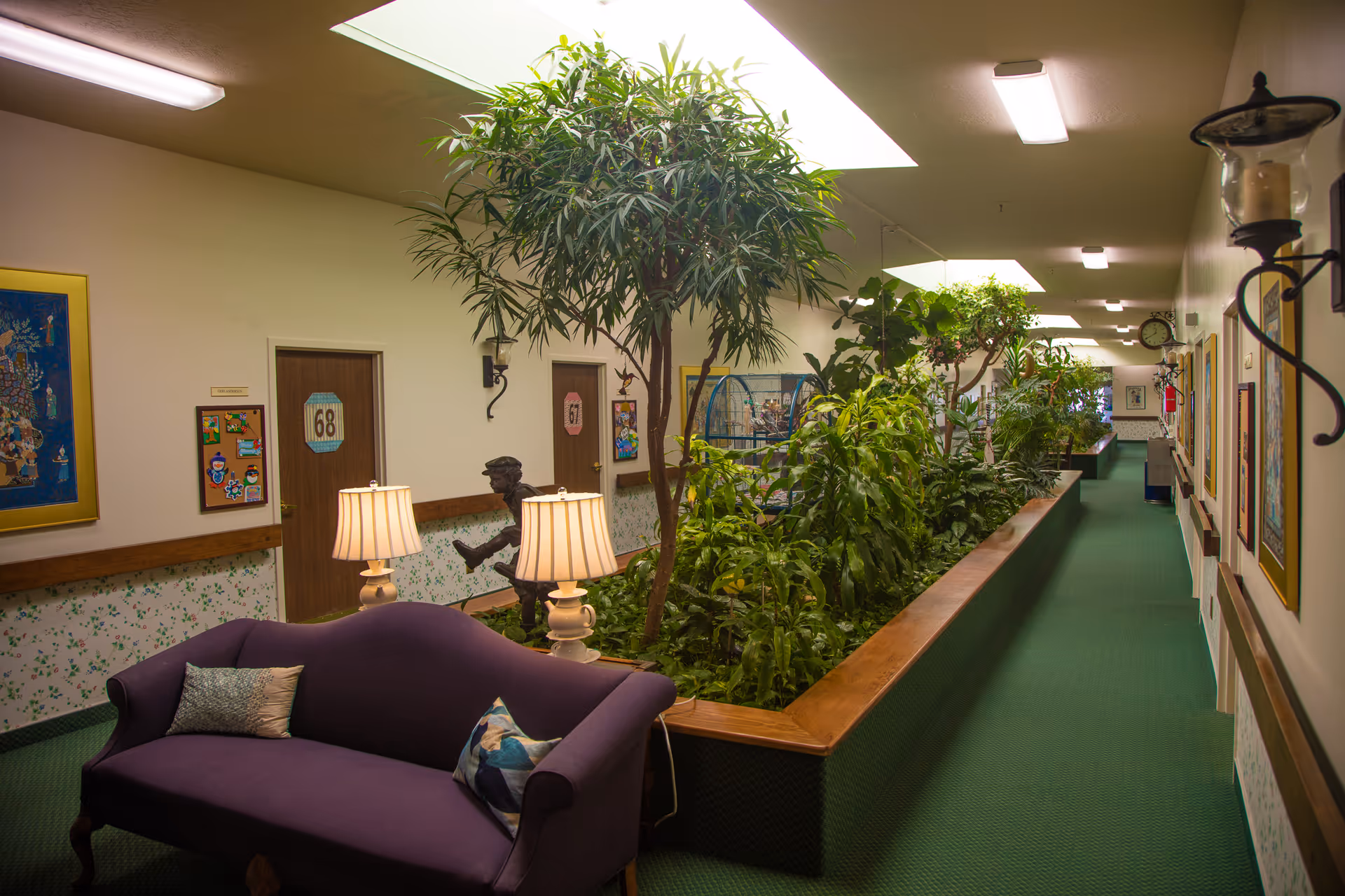 Indoor hallway of Gardens Assisted Living with green carpet and a large planter filled with various green plants and small trees under skylights. There is a purple couch with pillows and two table lamps in the foreground, doors numbered 67 and 68 on the left wall, framed artwork on the walls, and a bronze statue of a child sitting on the edge of the planter.