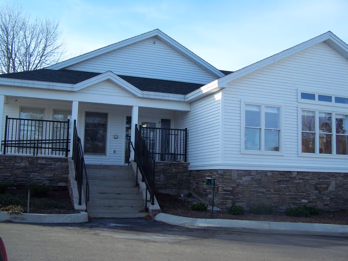 Front exterior of a white single-story building with stone foundation, steps leading to a covered porch and black railings.