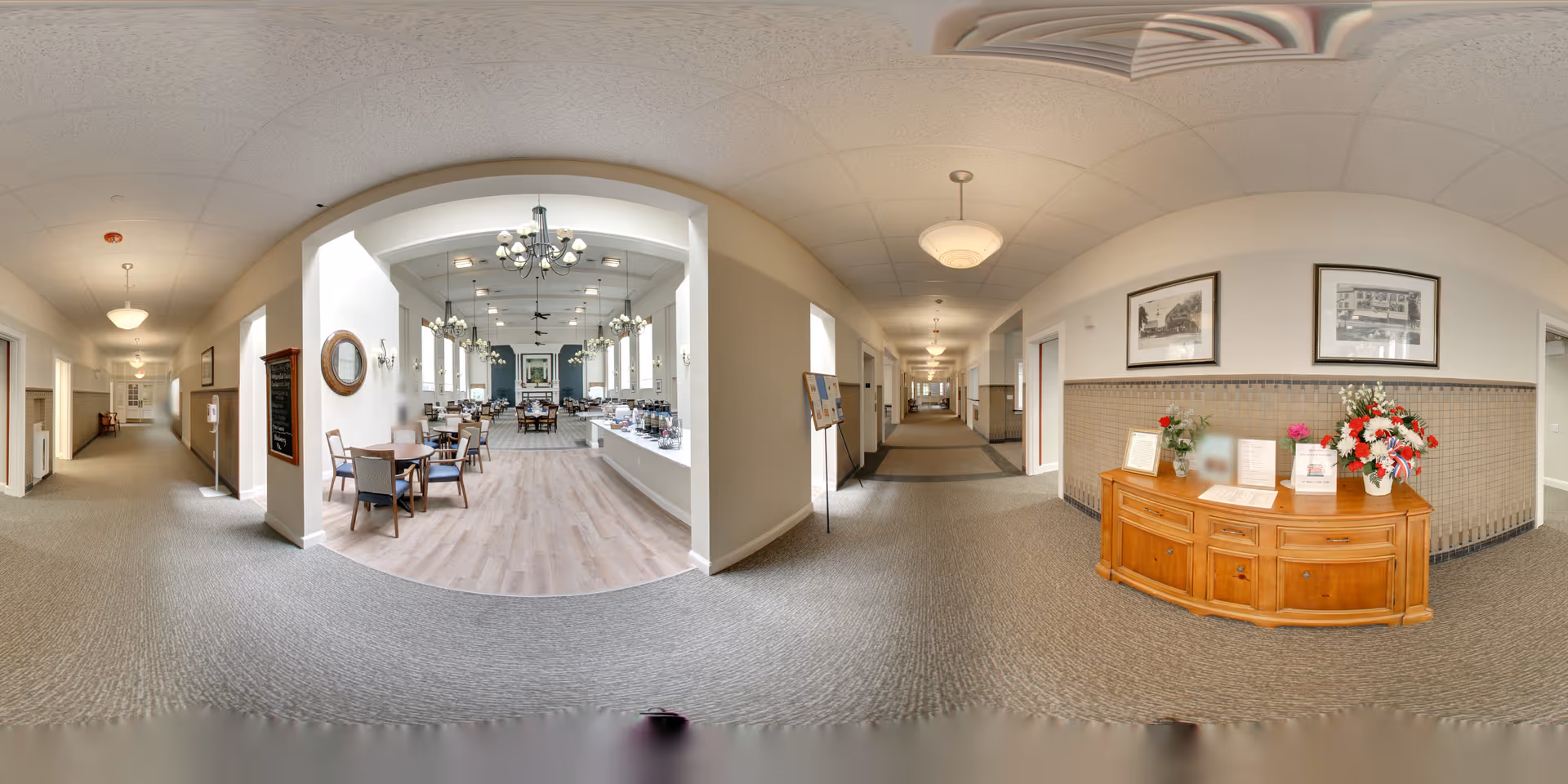 Panoramic view of an interior hallway in a senior living facility with carpeted floors and beige walls. To the left, there is a dining area with wooden tables and chairs, chandeliers, and large windows letting in natural light. To the right, there is a wooden sideboard with flower arrangements and framed pictures on the wall above it. The hallway extends into the distance with doors and light fixtures along the walls.