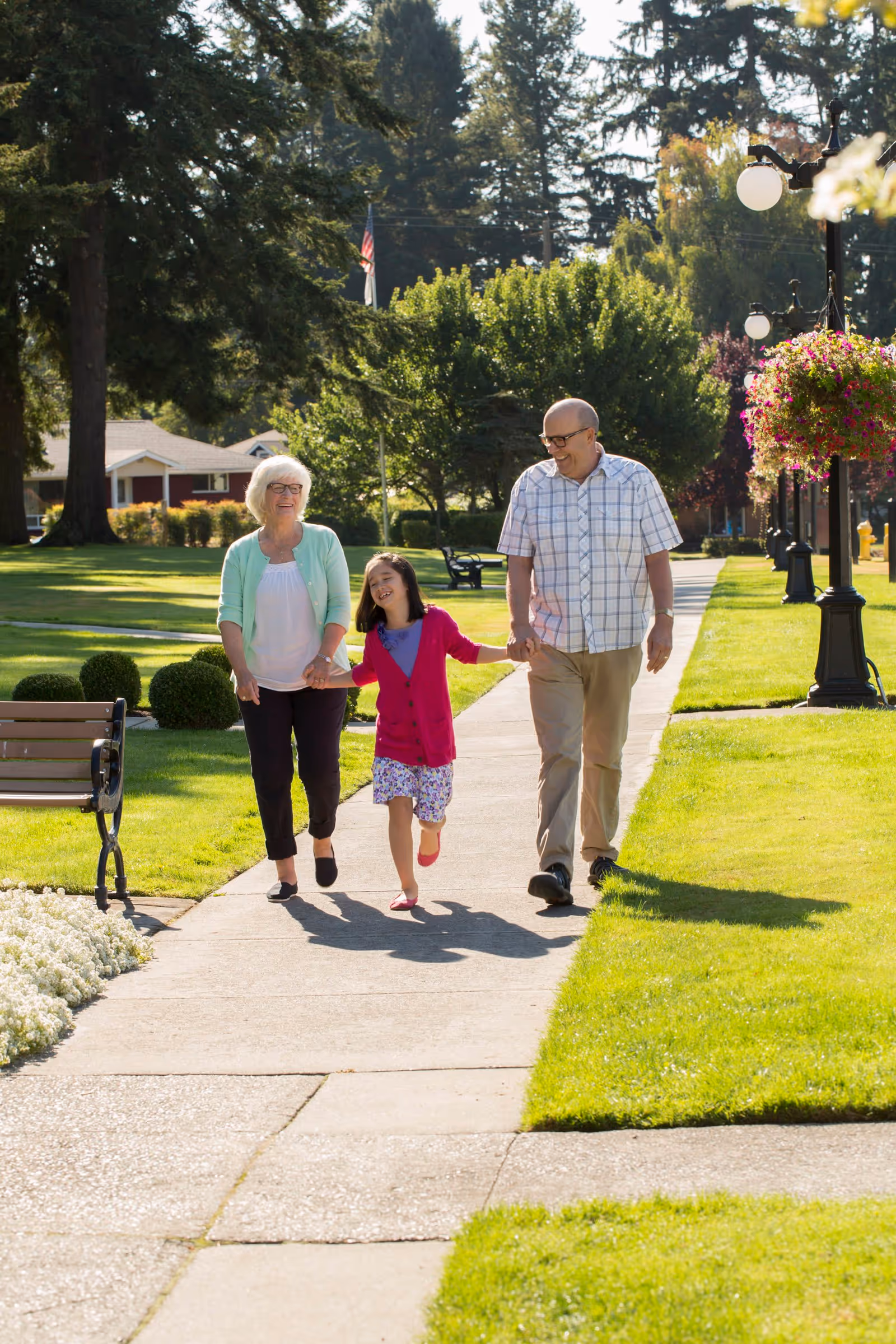 An older man and woman walk with a smiling young girl, holding hands along a sunny grassy sidewalk lined with benches and lamp posts.