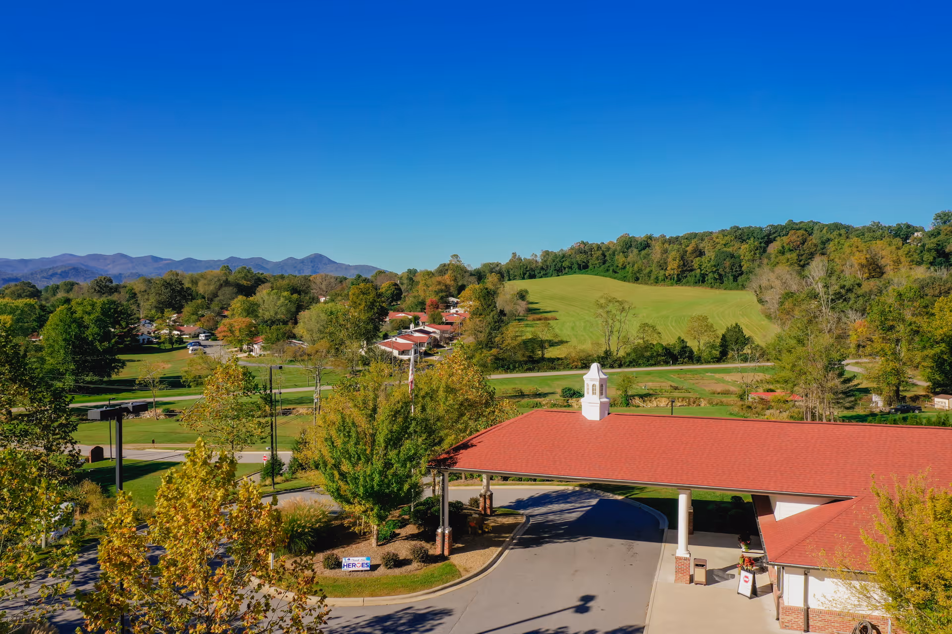 Aerial view of Pisgah Valley Retirement Community showing a covered entrance with a red roof and white cupola, surrounded by trees and greenery. In the background, there are rolling hills and mountains under a clear blue sky.