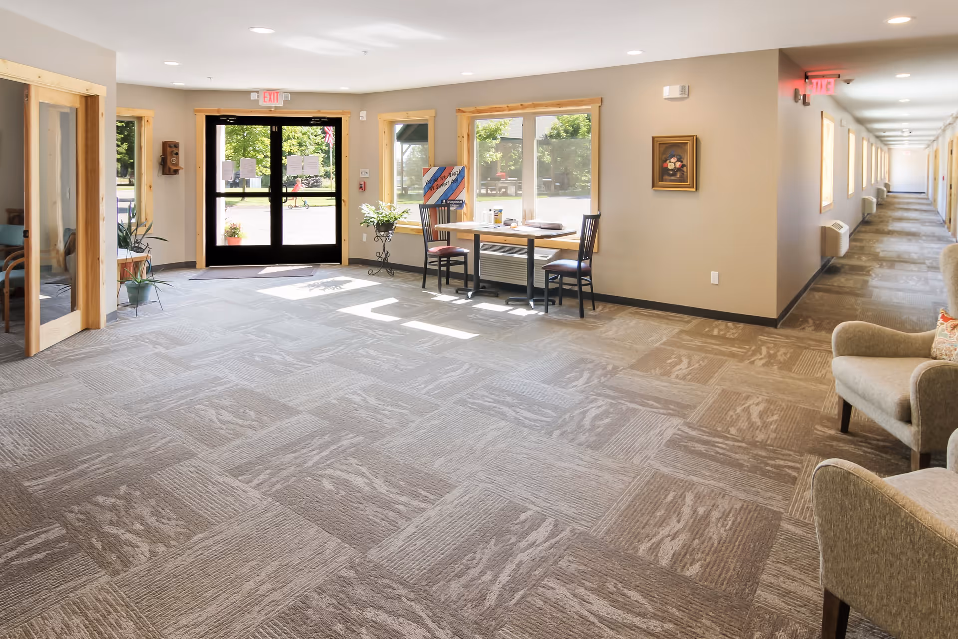 Bright and spacious hallway area in South Torch Assisted Living with large windows letting in natural light, a table with two chairs near the window, beige walls, carpeted floor, and armchairs along the right side. The hallway extends to the right with multiple doors and exit signs visible.