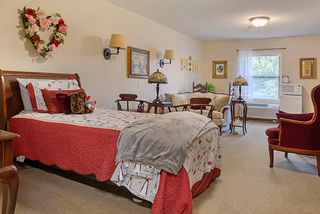 A bright senior living bedroom with a made bed, floral decor, a seating area with sofa and chairs, lamps, and a window.