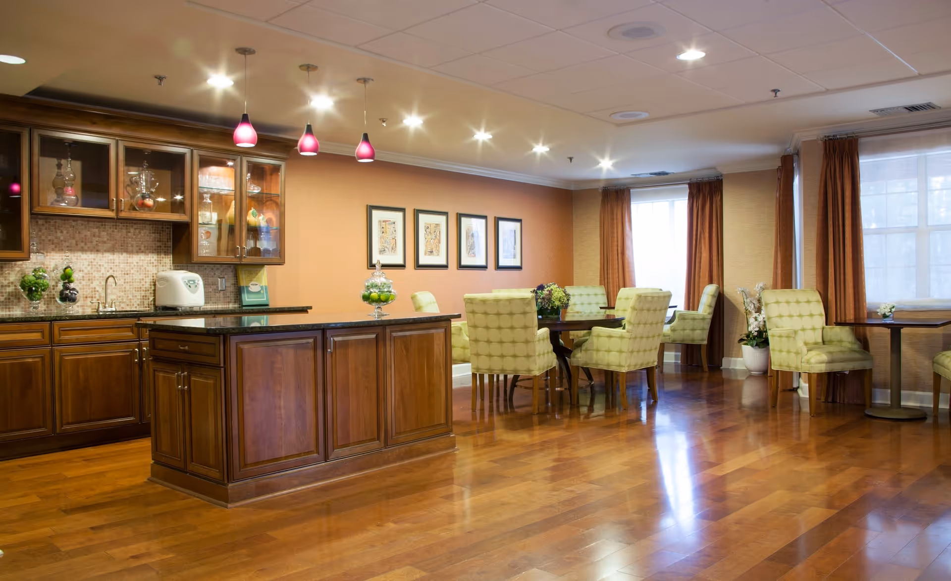 A spacious dining area with wooden flooring and warm lighting. The room features a kitchen island with dark granite countertop and wooden cabinets. There are several green cushioned chairs around dark wooden tables. The walls are painted in a warm beige tone with framed artwork, and large windows with brown curtains allow natural light to enter the room.