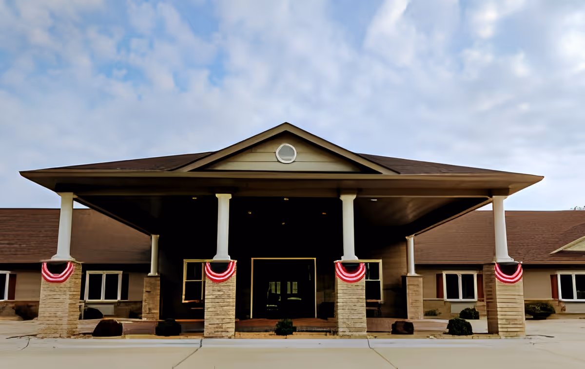 Front exterior view of a single-story building with a covered entrance supported by four columns wrapped with red, white, and blue bunting. The building has a brown roof and beige walls with several windows. The sky is partly cloudy.