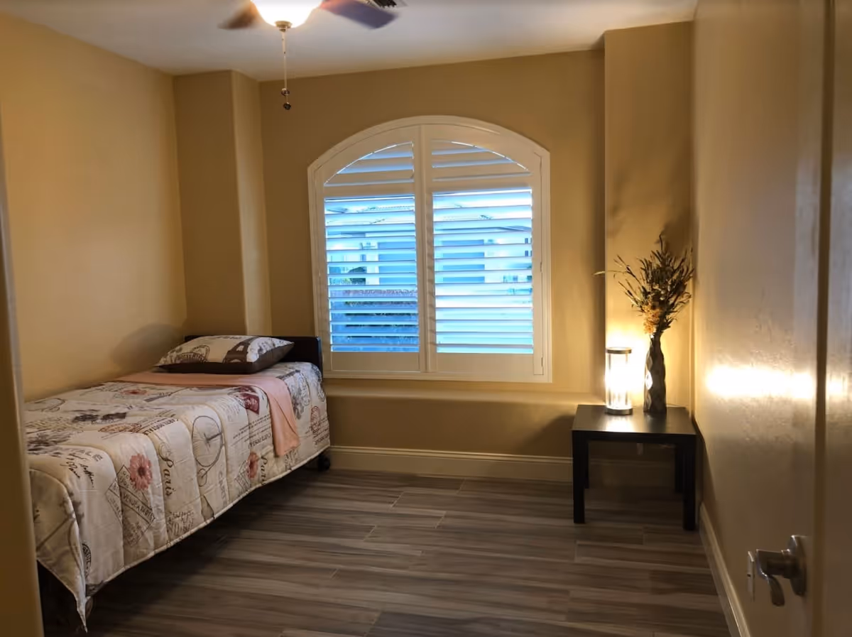 A small bedroom with a single bed covered in a patterned quilt and a pillow. There is a window with white plantation shutters letting in natural light. Next to the bed is a black side table with a lit lamp and a vase with flowers. The room has beige walls and wood-look tile flooring.