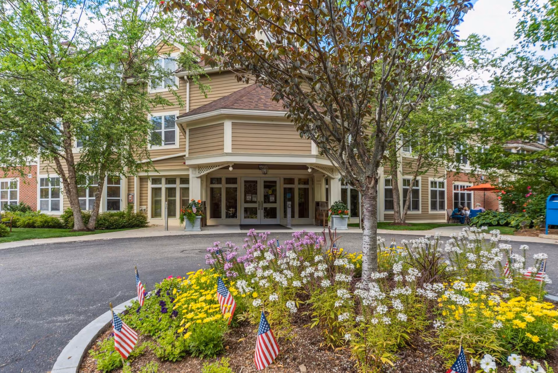 Front entrance of The Gables at Winchester senior living facility with a circular driveway, landscaped flower bed featuring yellow, white, and purple flowers, small American flags, and trees surrounding the building.