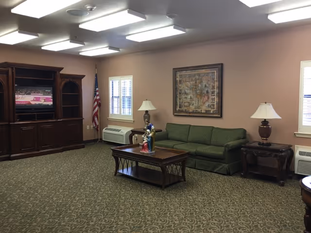 A communal living room with a green sofa, wooden coffee table, TV cabinet, lamps, framed artwork, and an American flag.