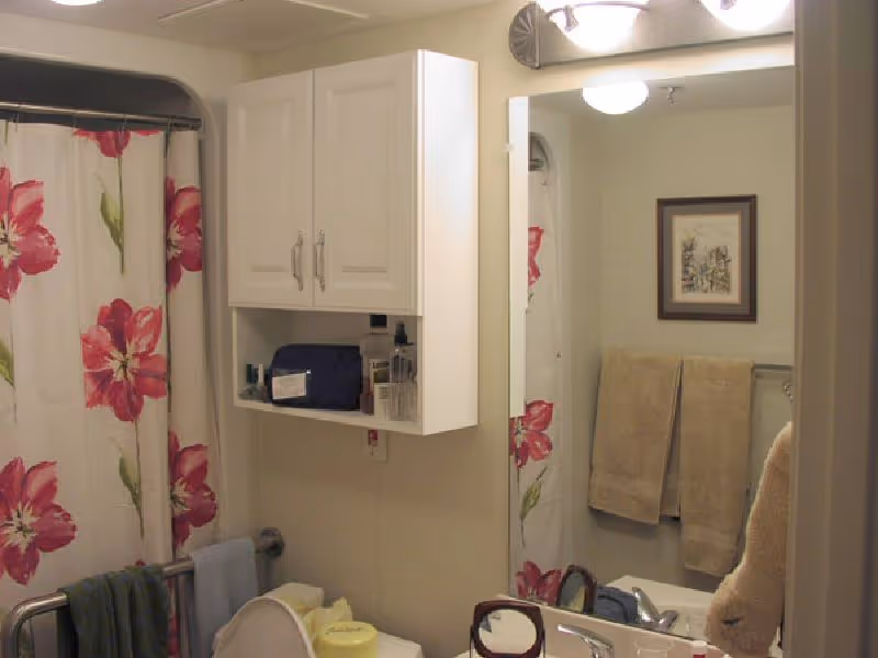 Bathroom with a floral shower curtain featuring large pink flowers, a white wall-mounted cabinet above the toilet, beige towels hanging on a towel rack, a framed picture on the wall, and a mirror reflecting part of the room.