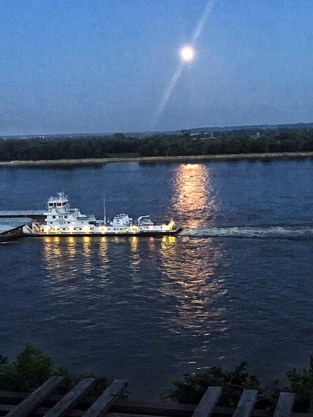 A nighttime view of a river with a brightly lit boat moving across the water. The full moon is visible in the sky, casting a reflection on the river's surface. Trees and land are visible in the background.