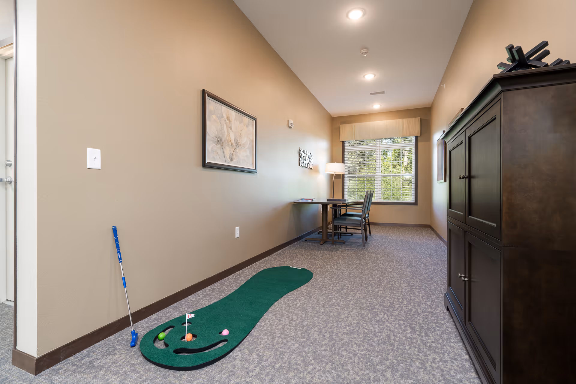 A narrow room with beige walls and carpeted floor featuring a small indoor putting green with golf balls and a putter. At the far end of the room, there is a table with two chairs next to a window with blinds and a floor lamp. A dark wooden cabinet is on the right side, and a framed floral artwork hangs on the left wall.