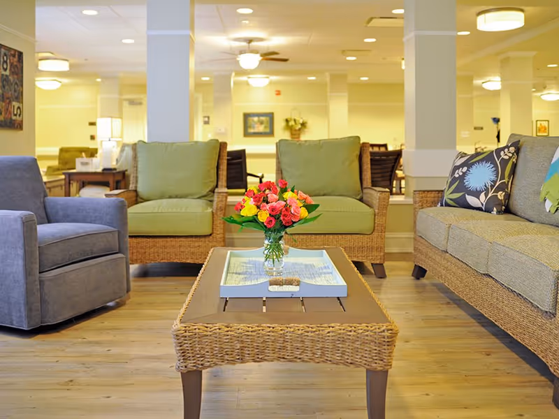 A bright senior living lounge with wicker sofas and armchairs arranged around a coffee table holding a vase of colorful flowers.