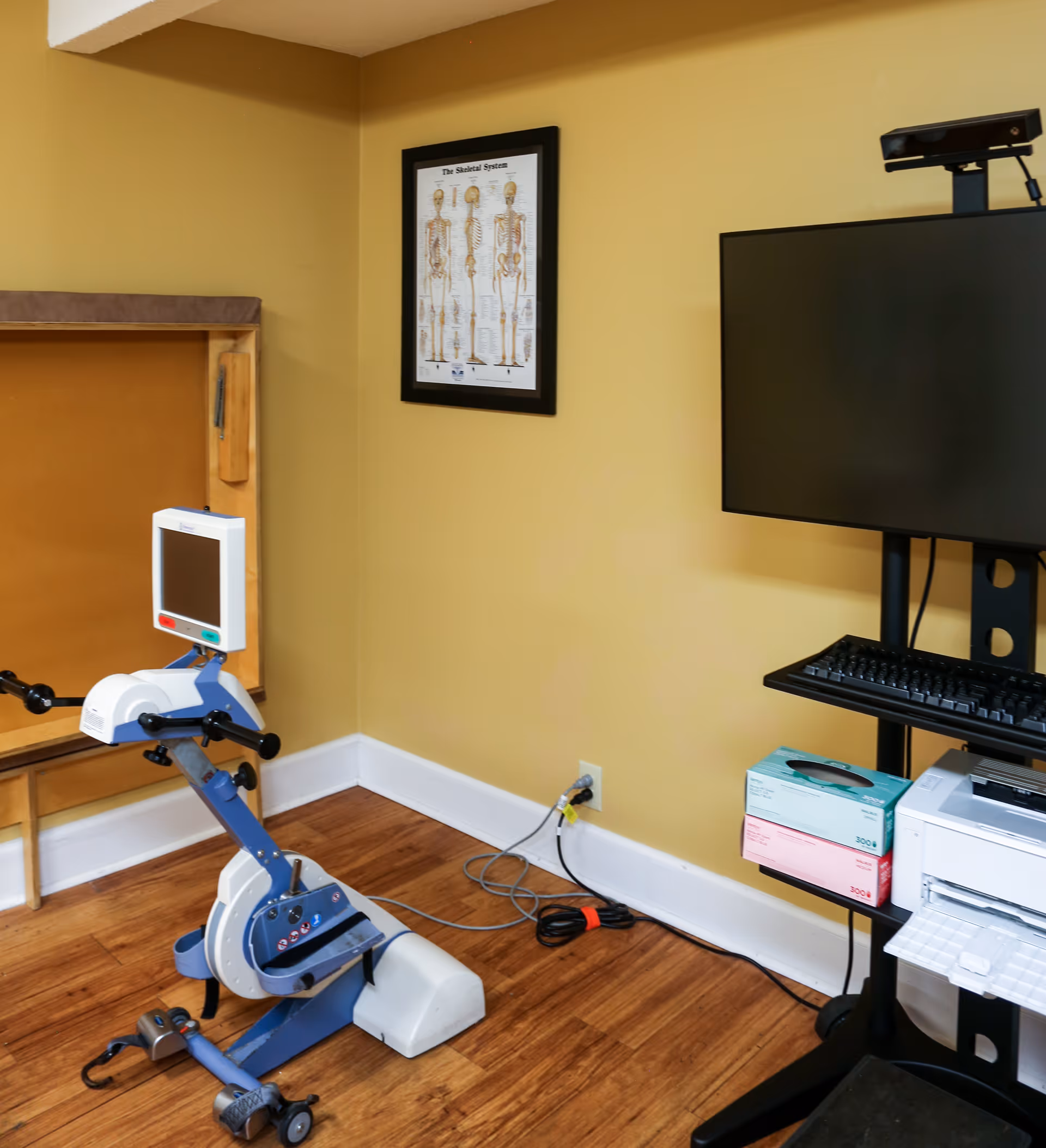 A corner of a room with a small exercise machine featuring a digital screen, a framed anatomical poster of the skeletal system on a yellow wall, and a black stand holding a computer monitor, keyboard, printer, and boxes of medical gloves on a wooden floor.