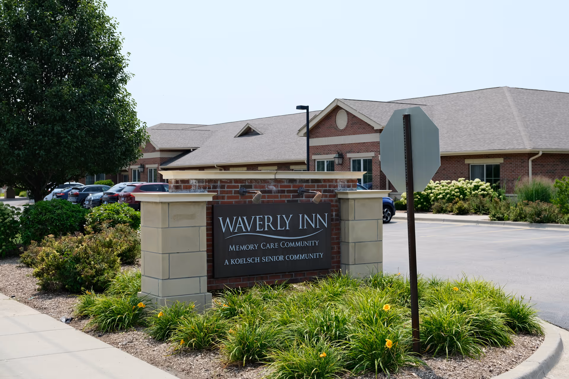 Exterior view of Waverly Inn Memory Care Community building with a brick and stone sign in front surrounded by green plants and yellow flowers. Several parked cars and a tree are visible in the background.