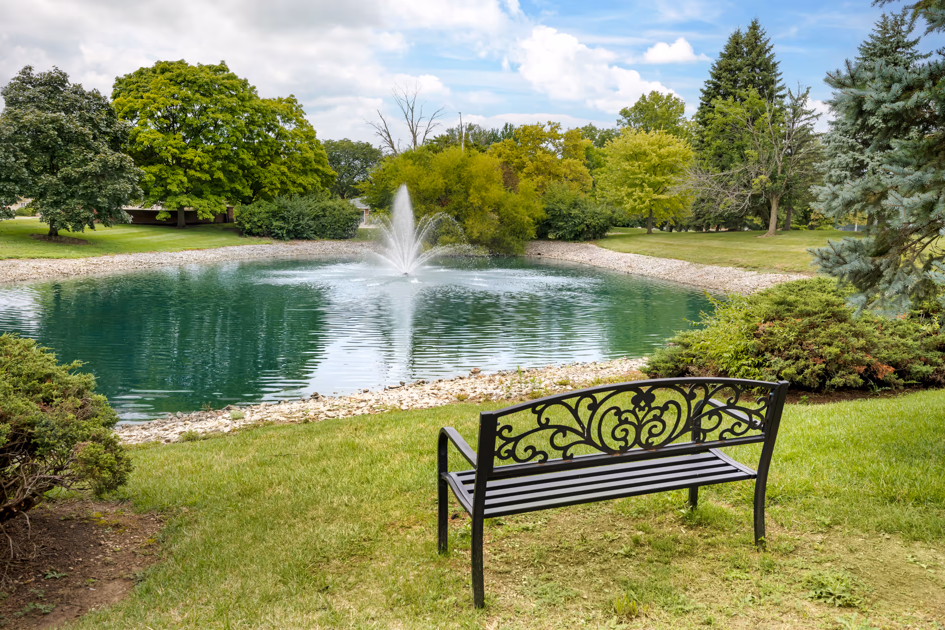 A black decorative metal bench on a grassy area facing a small pond with a water fountain in the center. The pond is surrounded by rocks and lush green trees and bushes under a partly cloudy sky.