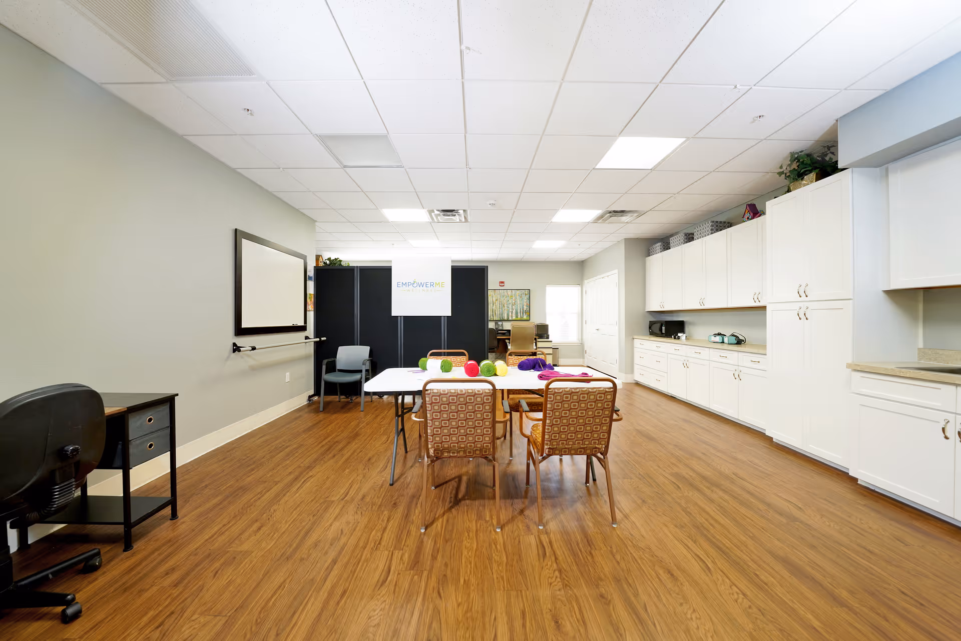 A spacious, well-lit activity room with wood flooring and a white ceiling. There is a table in the center with four patterned chairs around it, and colorful yarn balls on the table. The right side of the room features white cabinets and countertops, while the left side has a black desk and chair. At the back, there is a black partition with a sign that reads 'EMPOWER ME'.