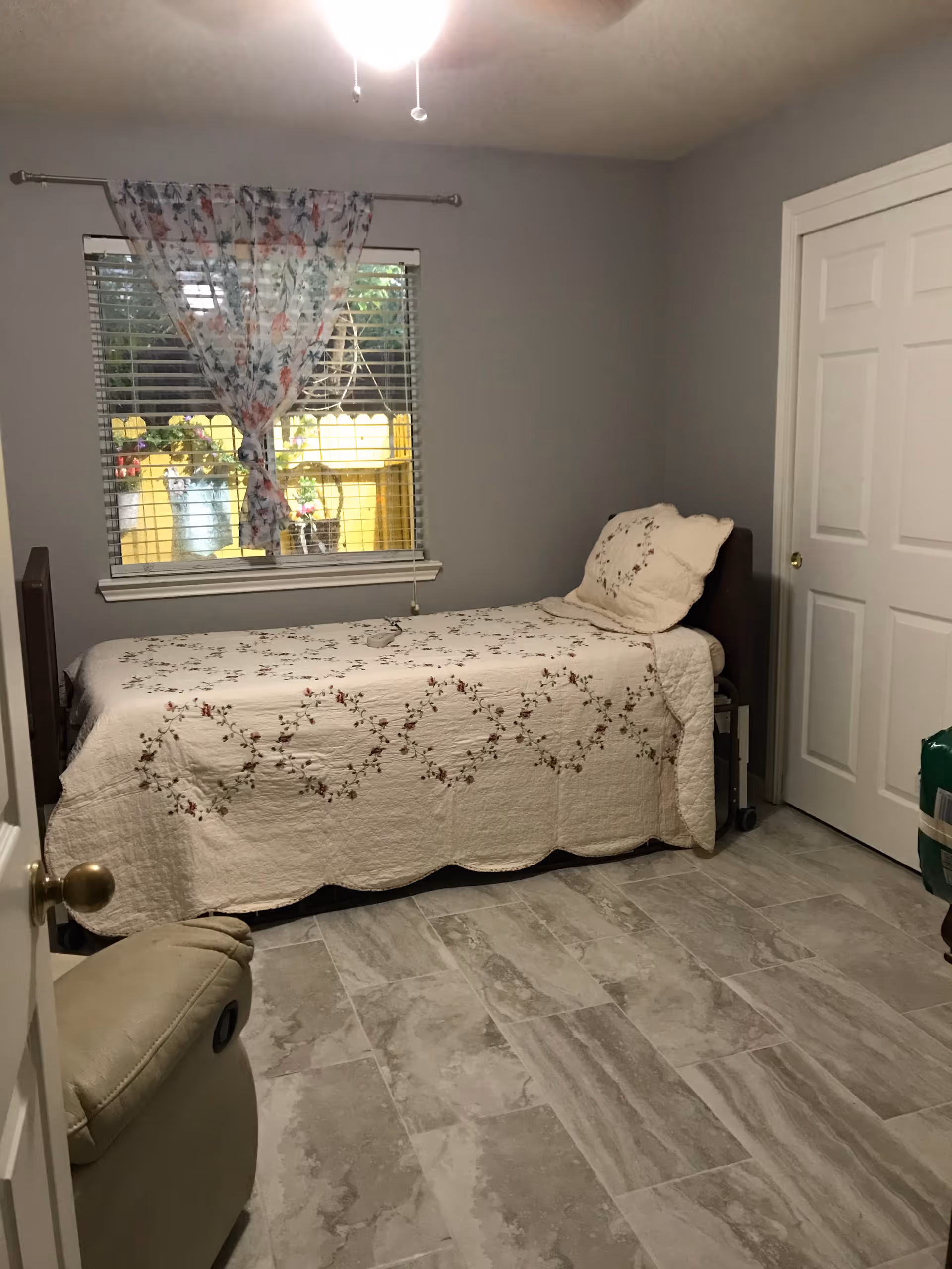 A small bedroom with a single bed covered with a cream-colored quilt featuring a floral pattern. There is a matching pillow on the bed. A window with floral curtains tied in the middle is above the bed, showing a yellow fence and some plants outside. The room has light gray walls, a tiled floor, a closed white door, and a beige recliner chair partially visible in the foreground.