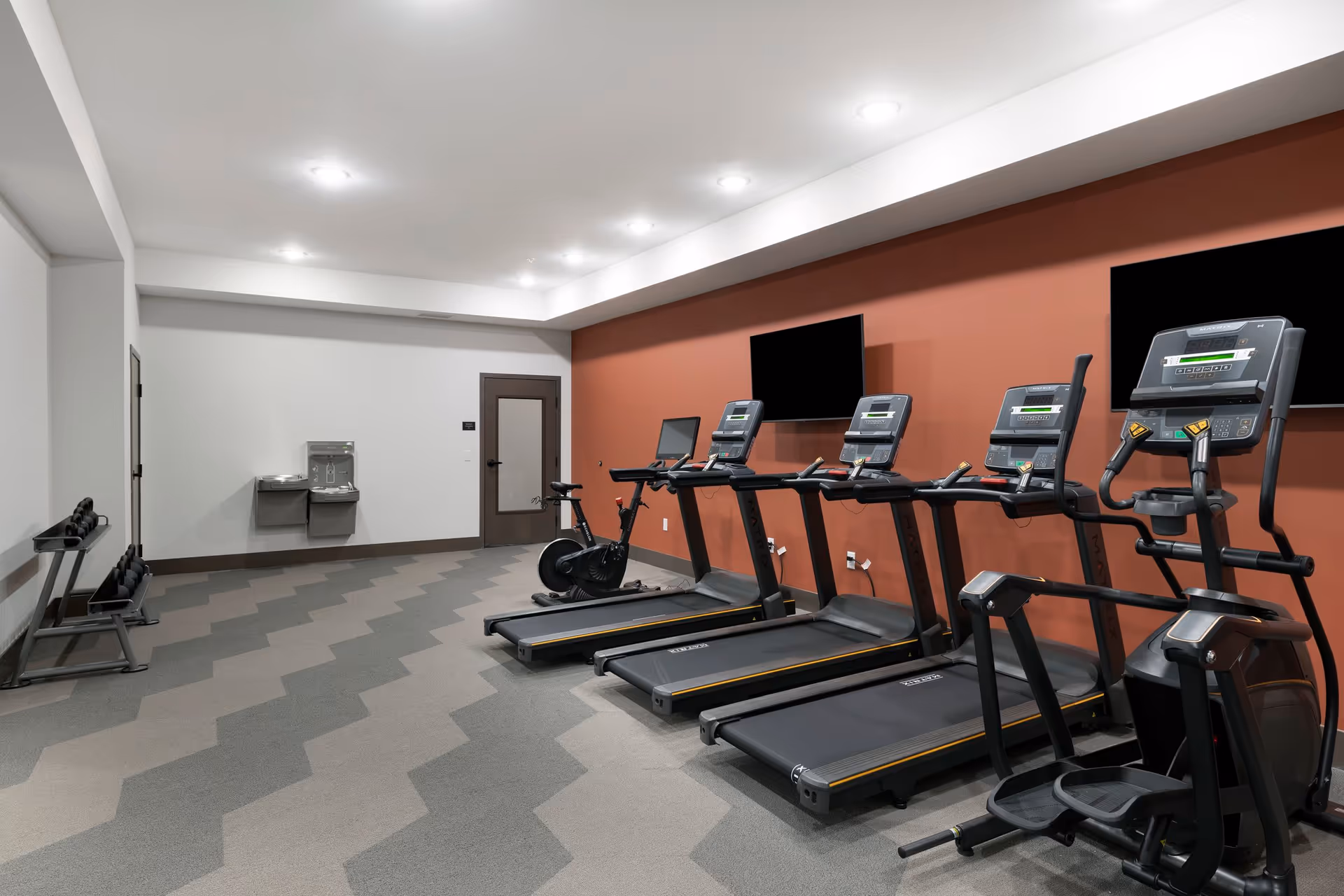 A fitness room with four treadmills, an elliptical machine, a stationary bike, and a rack of dumbbells. The room has a patterned carpet floor, white ceiling with recessed lighting, and a red accent wall with two mounted flat-screen TVs. There is a water fountain and a door on the far wall.
