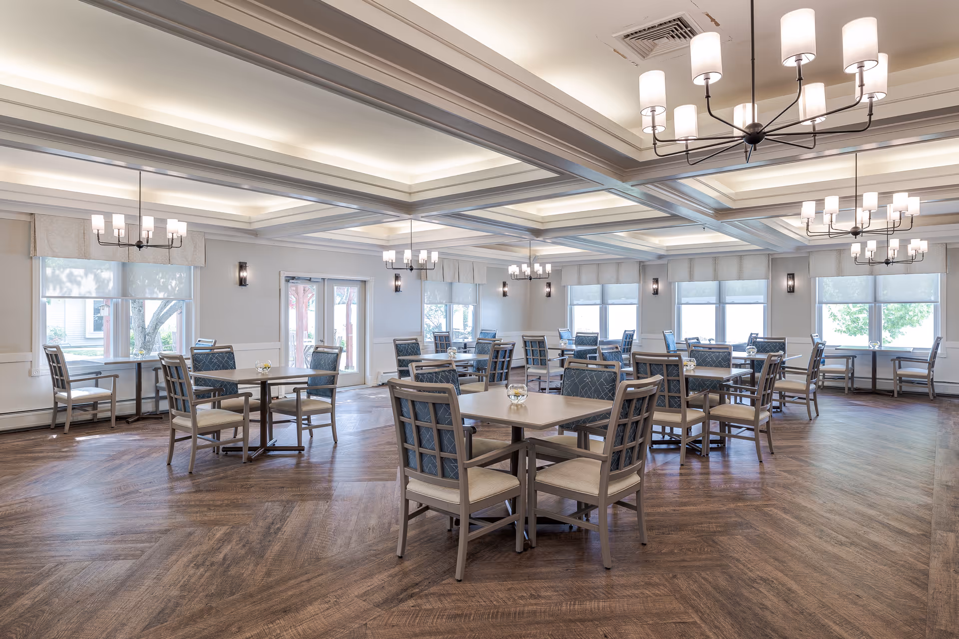 A spacious dining room with multiple square tables and chairs arranged neatly. The room features large windows with white shades allowing natural light to brighten the space. The ceiling has recessed lighting and several modern chandeliers with cylindrical lampshades. The floor has a wood pattern, and the walls are painted light gray with white trim.