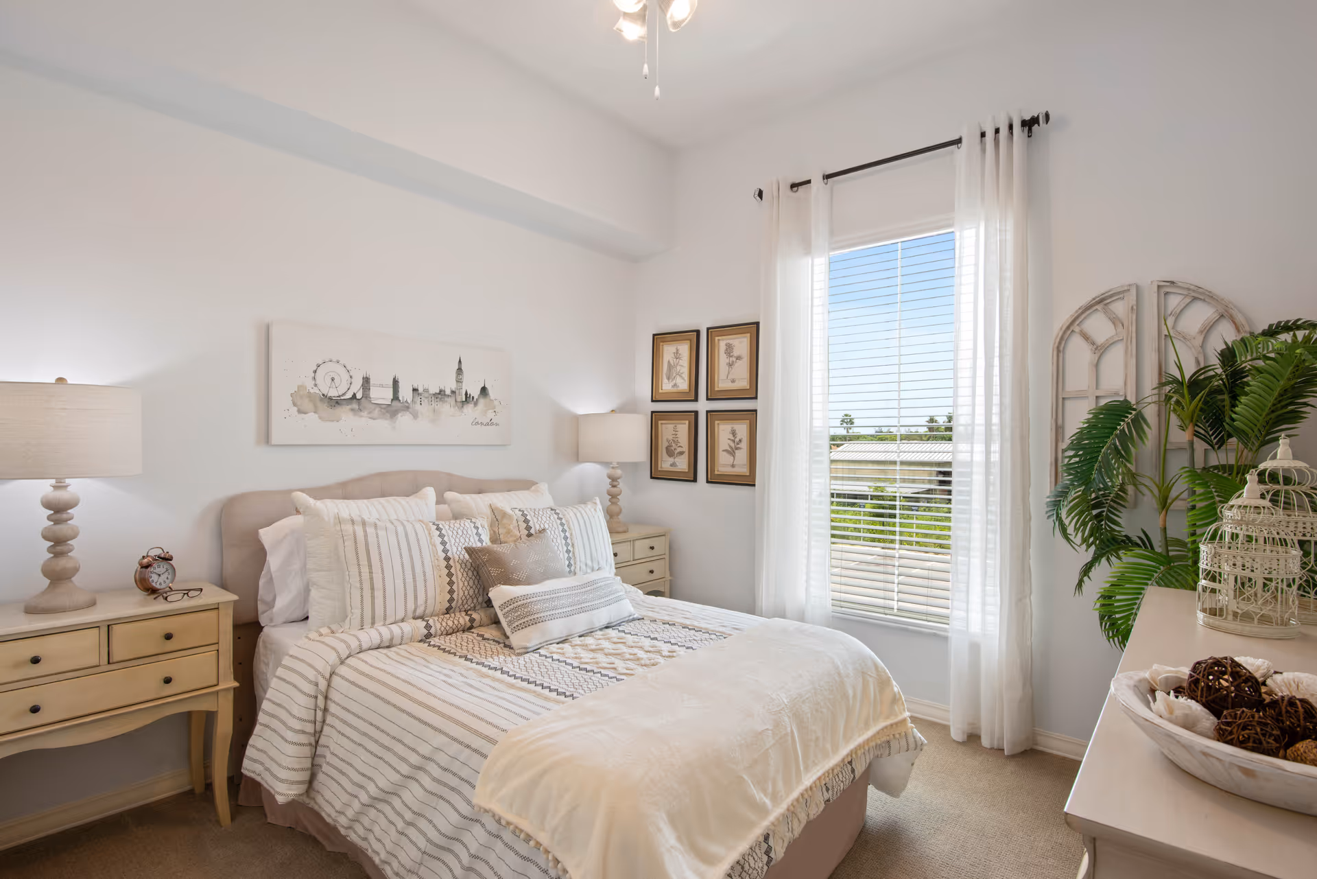 A bright and cozy bedroom with a neatly made bed featuring multiple pillows and a striped comforter. Two matching bedside tables with lamps flank the bed. A window with sheer white curtains lets in natural light, and decorative framed pictures hang on the wall. A dresser with decorative items and a green plant is visible on the right side of the room.