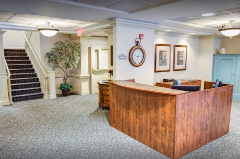 A carpeted lobby and reception area with a wooden front desk, staircase, potted plant, wall clock and framed artwork.