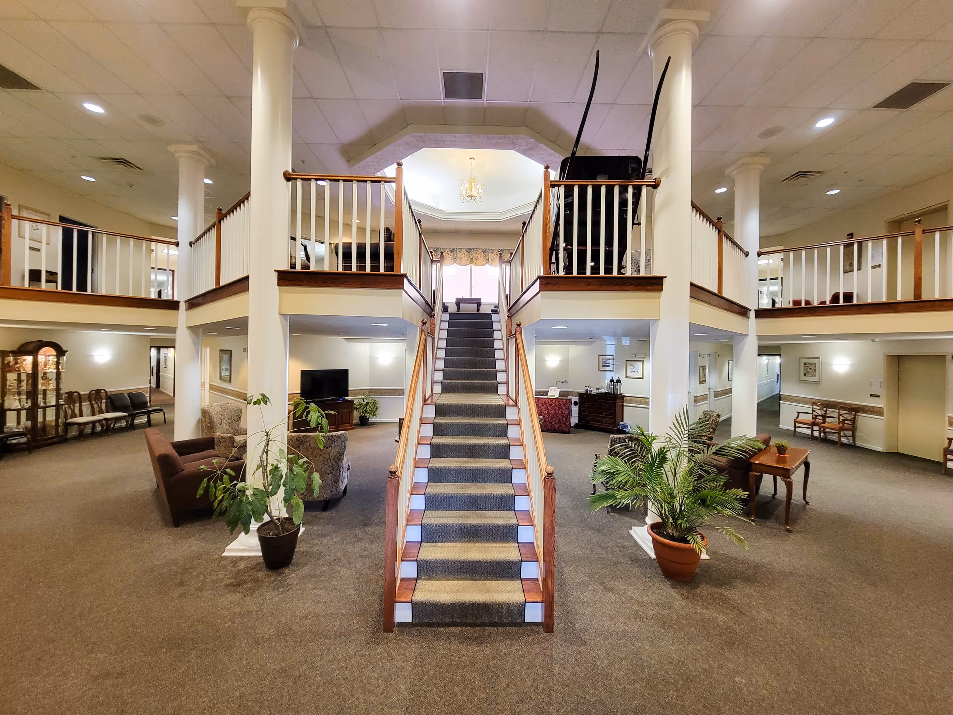 Interior view of a senior living facility lobby with a central carpeted staircase leading to an upper level. The space features white columns, wooden railings, seating areas with armchairs and sofas, potted plants, and soft lighting. There is a chandelier hanging from the ceiling above the staircase.