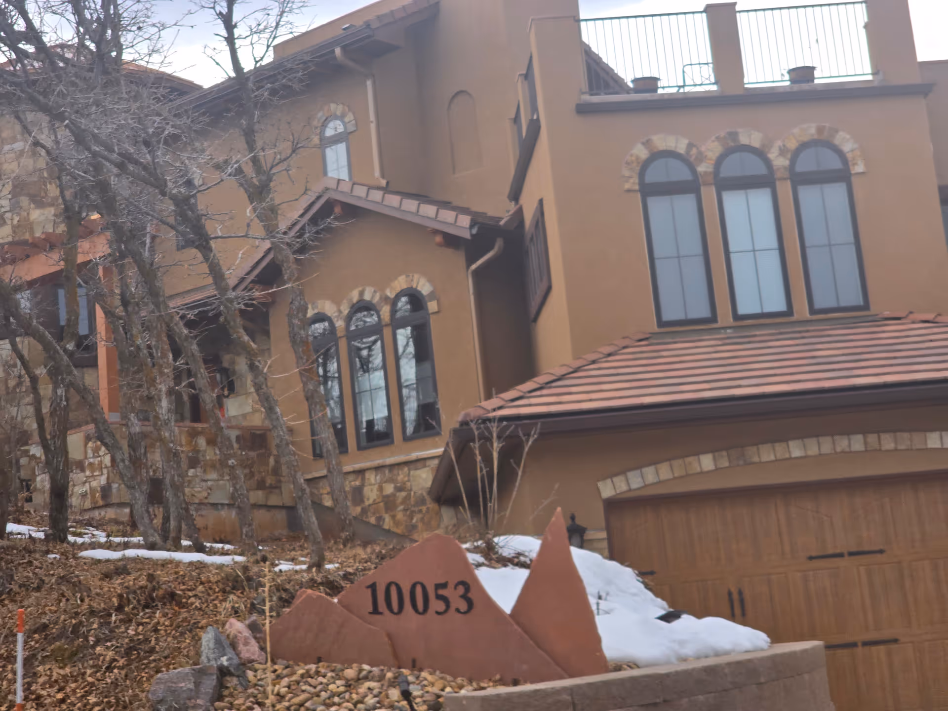 Exterior view of a large, multi-story building with stone and stucco walls, arched windows, and a tiled roof. In the foreground, there is a stone sign with the number 10053 on it, surrounded by rocks and some snow. Leafless trees are visible around the building.