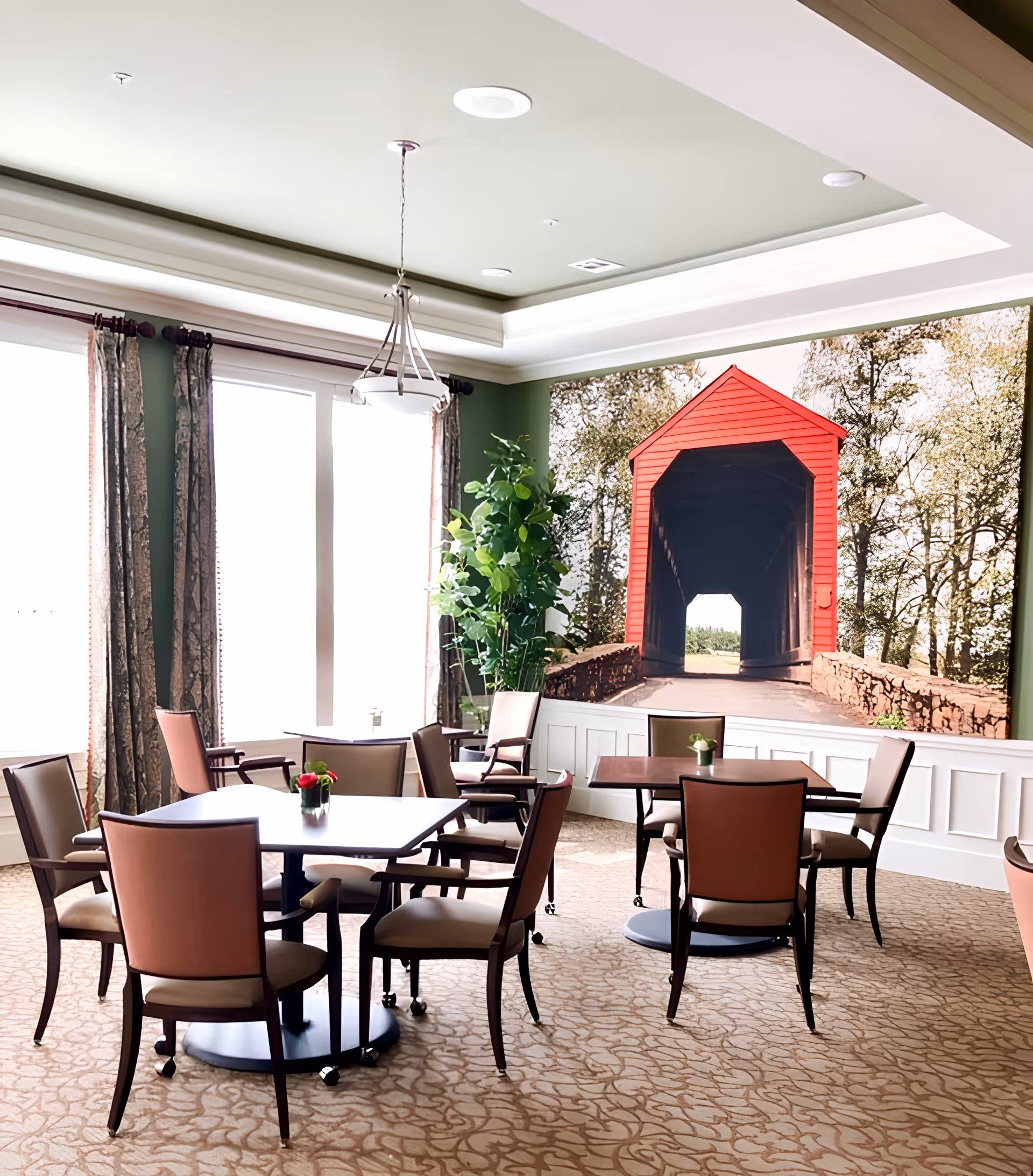 Bright dining room with several tables and chairs, a large mural of a red covered bridge on the wall, and tall windows with curtains.