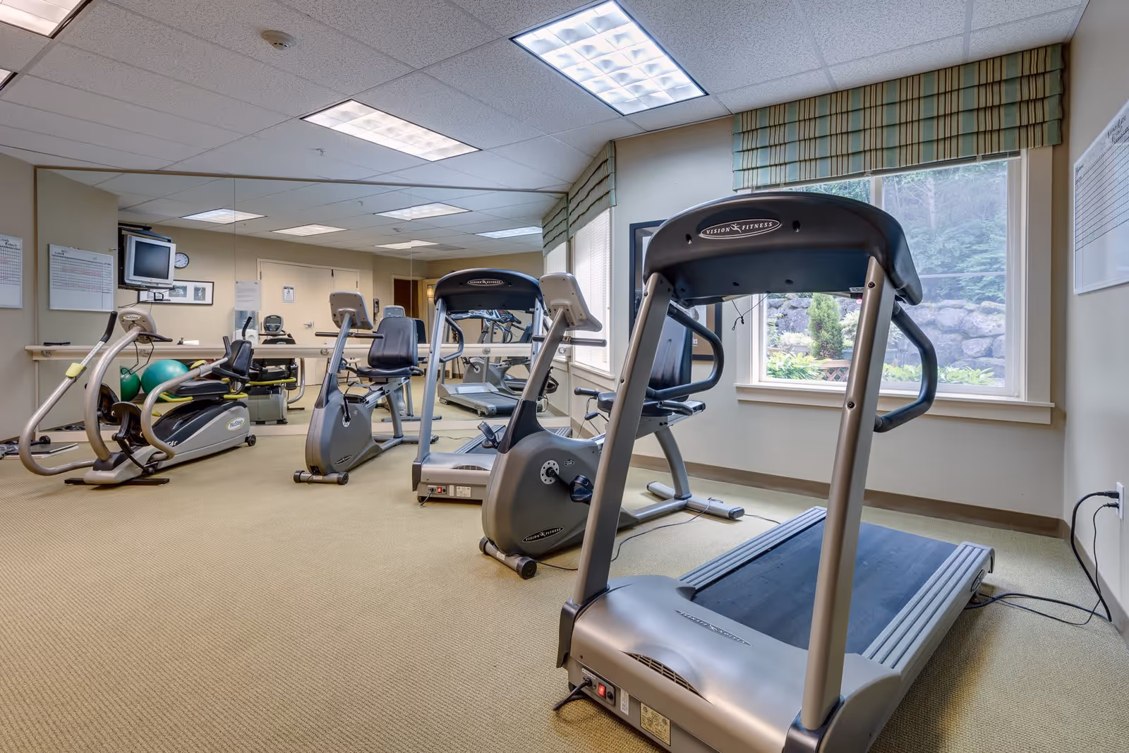 A fitness room with exercise equipment including a treadmill, stationary bikes, and an elliptical machine. The room has beige carpet, large windows with striped valances, and a wall covered with mirrors reflecting the equipment. There is a television mounted on the wall and some fitness balls in the corner.