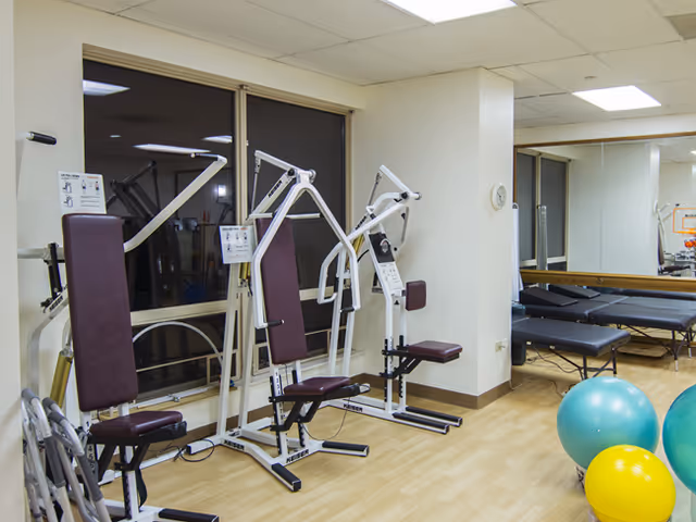 An exercise/therapy room with seated weight machines, treatment tables, mirrors, and colorful exercise balls.