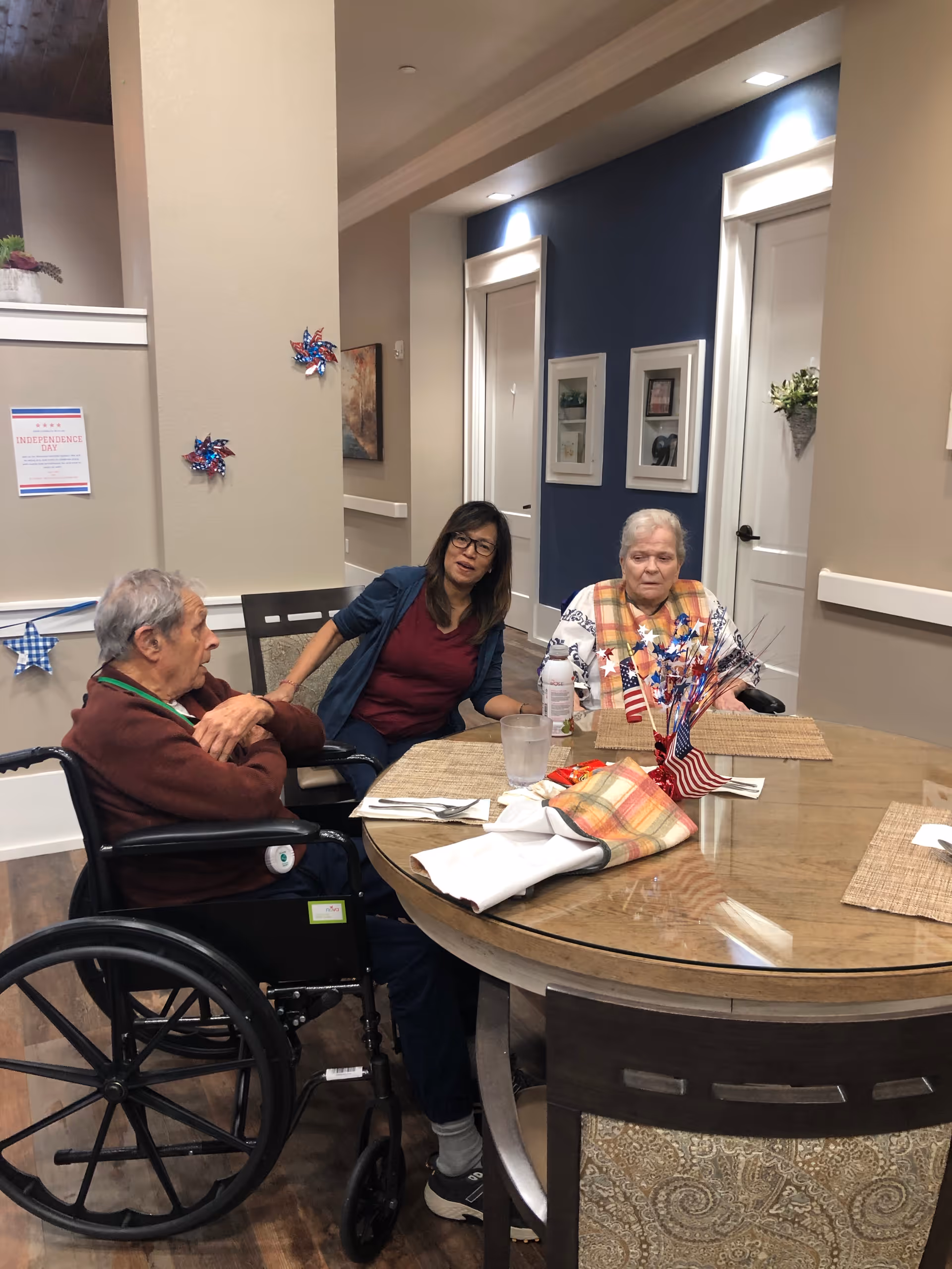 Three people sitting around a round dining table in a well-lit room. One person is in a wheelchair, another is a woman wearing glasses, and the third is an elderly woman with a plaid shawl. The table has placemats, napkins, a glass of water, and patriotic decorations including small American flags and red, white, and blue star-shaped ornaments. The room has beige walls with a dark blue accent wall and white doors in the background. There are framed pictures on the wall and some Independence Day decorations.