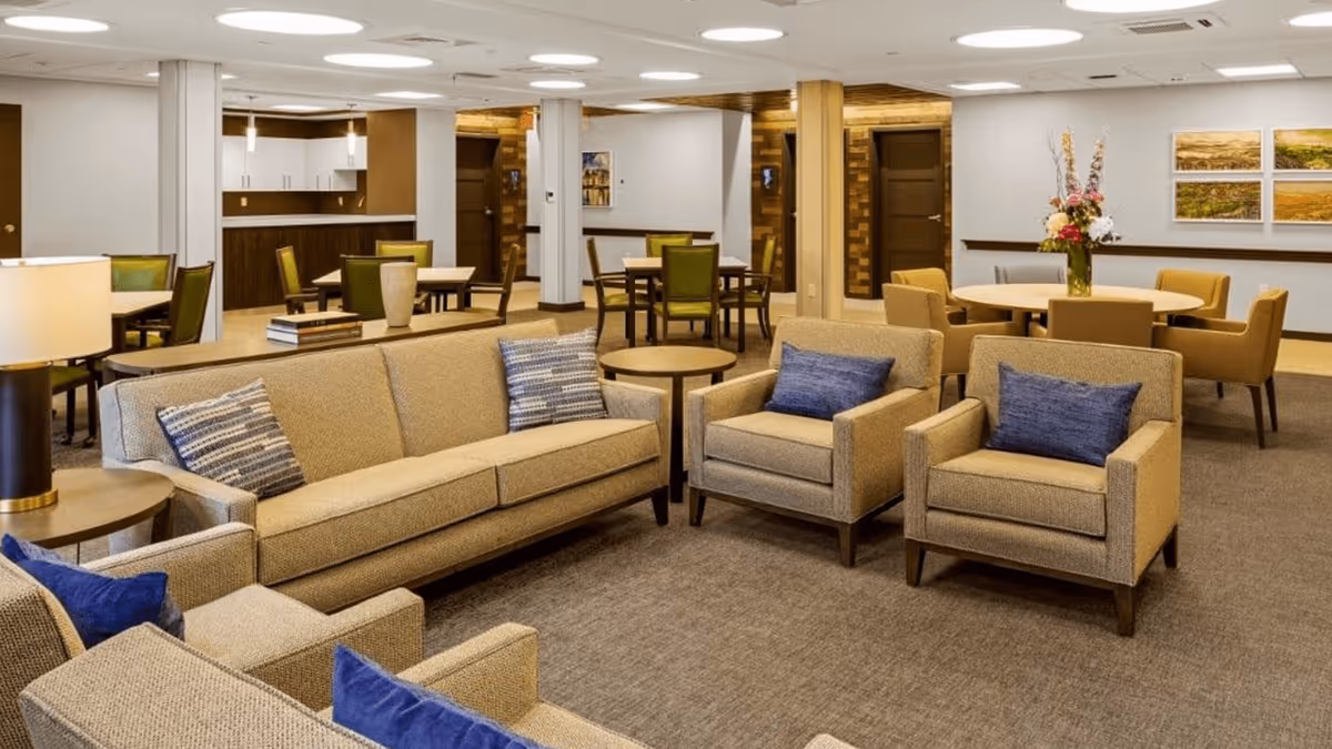 A spacious and well-lit common area in a senior living facility featuring beige upholstered sofas and armchairs with blue and patterned cushions, round wooden side tables, and several dining tables with green and beige chairs. The walls are decorated with framed landscape artwork and a vase with flowers is placed on one of the round tables. The ceiling has circular recessed lighting fixtures.