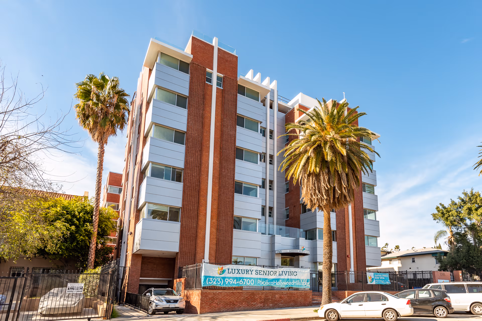 Front exterior of a multi-story brick-and-white senior living building with palm trees and parked cars.