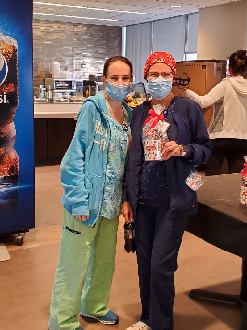 Two healthcare workers wearing scrubs and face masks stand together indoors. One is dressed in light green pants and a blue hoodie, while the other wears dark blue scrubs and a red bandana, holding a gift bag and a bottle of soda. Behind them is a Pepsi vending machine and a counter with food items.