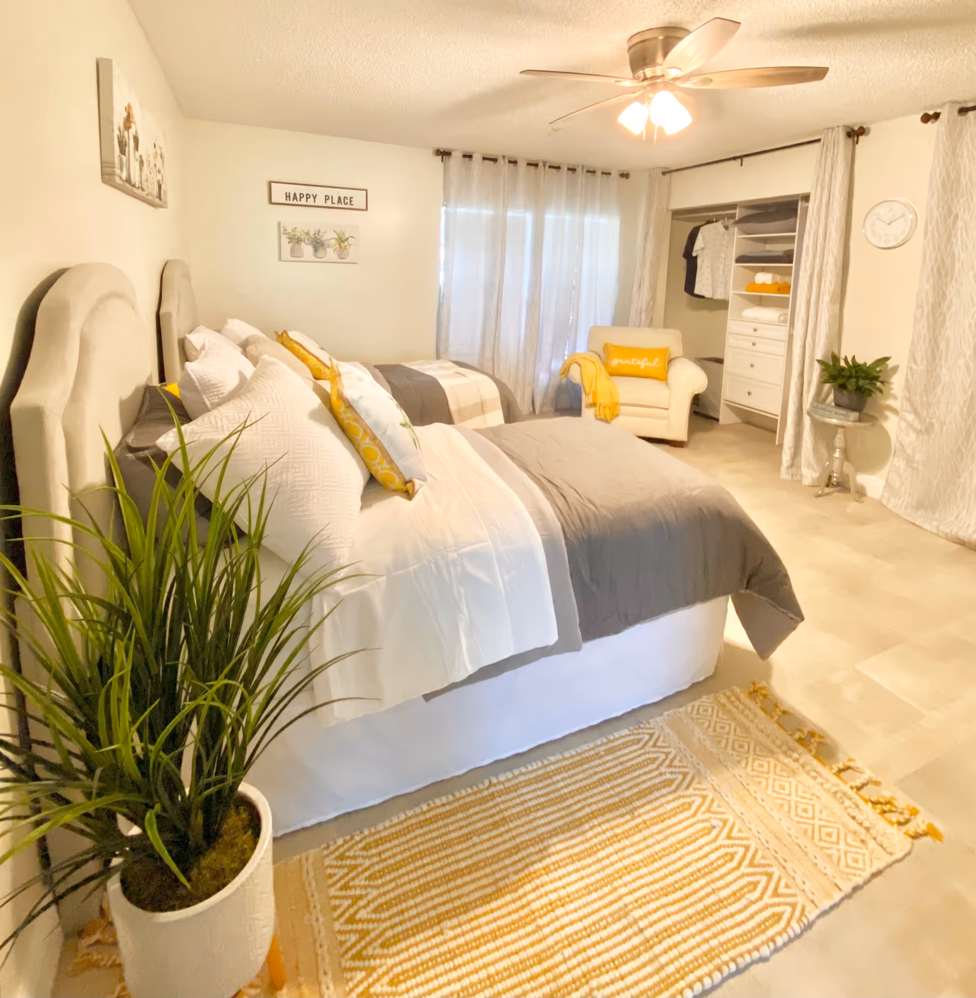 A bright and cozy bedroom with two beds featuring white and gray bedding accented with yellow pillows. The room has a ceiling fan with lights, a white armchair with a yellow pillow and throw, an open closet with shelves and hanging clothes, a small round table with a plant, a large potted plant near the beds, and light-colored curtains covering the windows. Wall decor includes a 'HAPPY PLACE' sign and framed pictures of plants.
