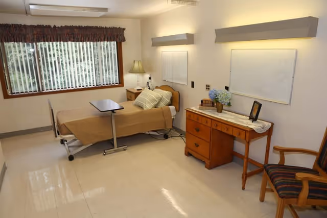 A simple, clean bedroom in a senior living facility with a single bed covered in a beige blanket, a bedside table with a lamp, a wooden dresser with a lace runner, a vase of flowers, and a framed picture. There is a large window with vertical blinds letting in natural light, a wooden chair with striped upholstery, and two whiteboards mounted on the wall.