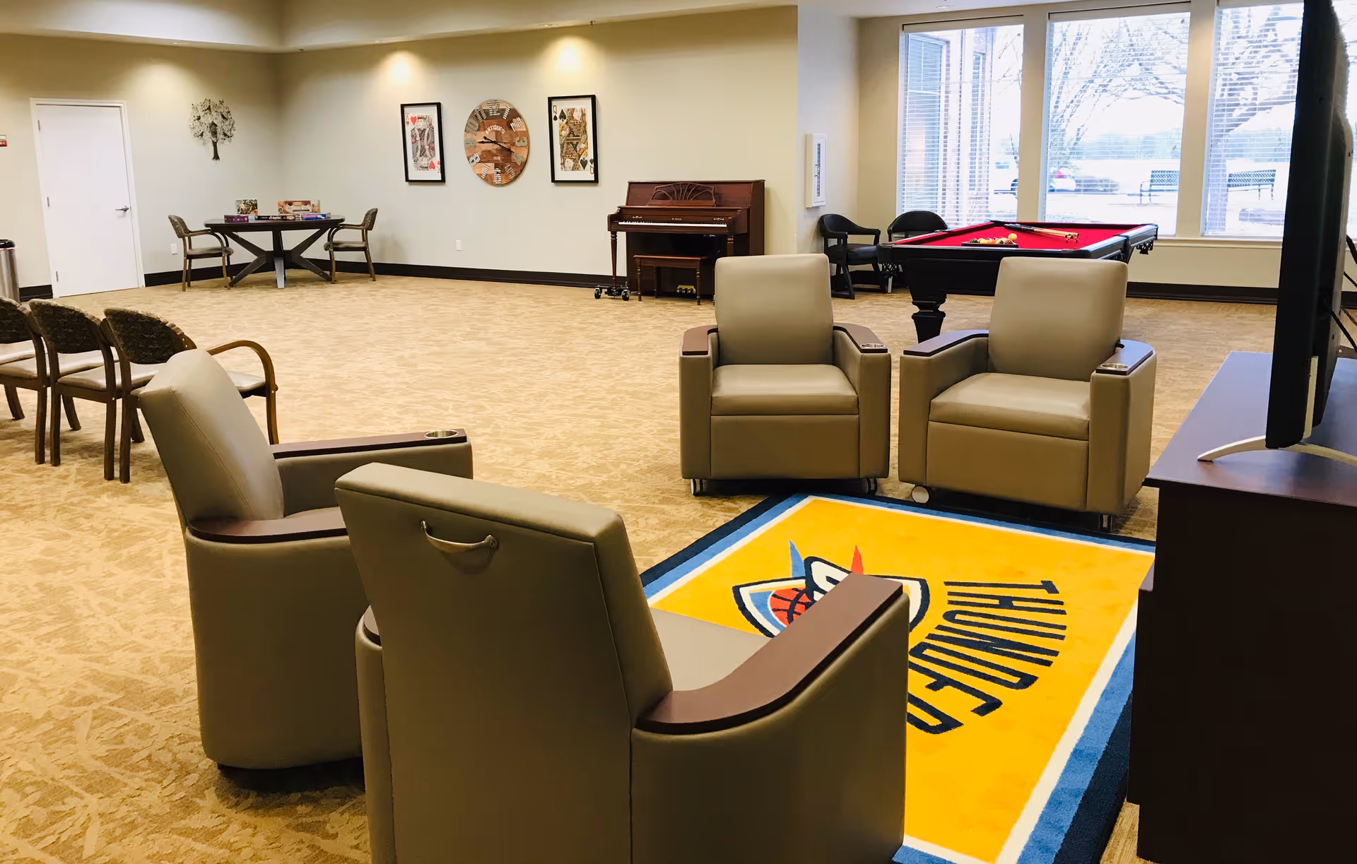 A spacious common room with beige carpet and neutral walls featuring four beige armchairs arranged around a TV on a stand. In the background, there is a pool table with a red felt top, a piano, a round table with chairs, and wall decorations including framed art and a clock. Large windows let in natural light.