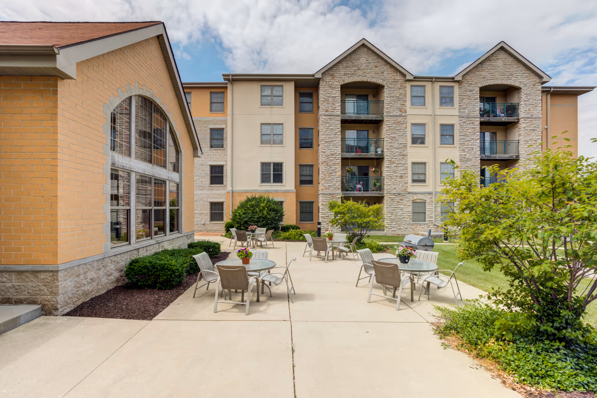 Outdoor patio area with several round tables and chairs arranged on a concrete surface. Each table has a small flower pot with colorful flowers. The patio is surrounded by green bushes and trees, with a multi-story building featuring balconies and large windows in the background under a partly cloudy sky.