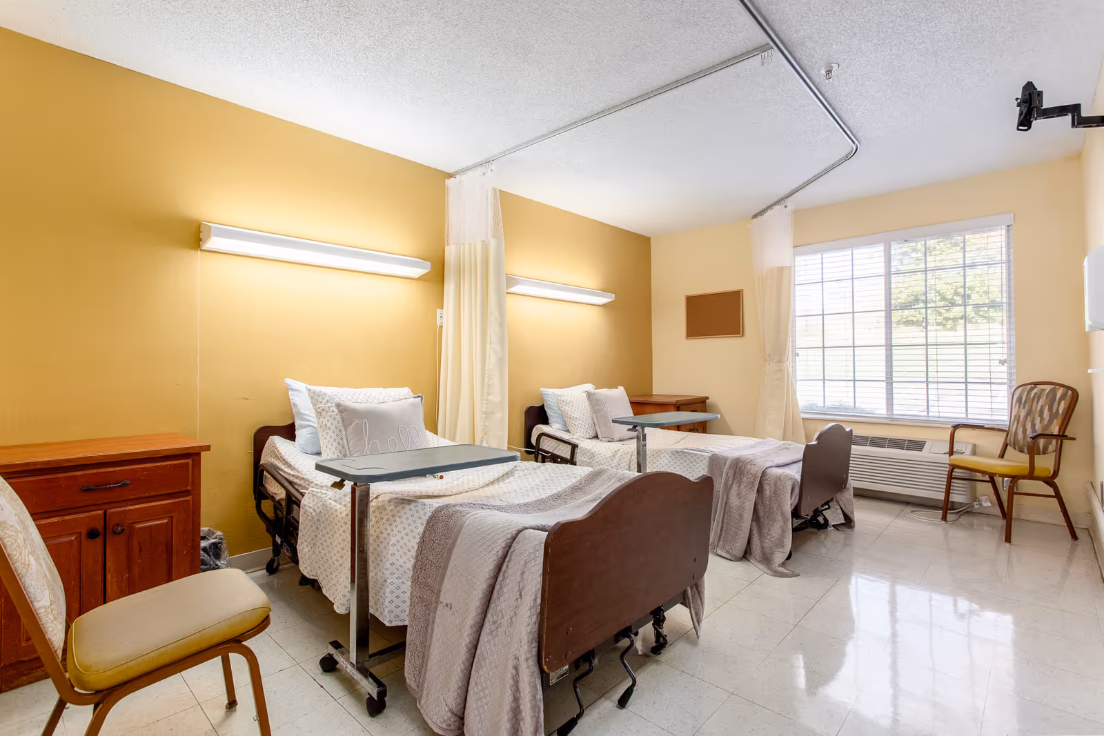Two hospital-style beds in a bright nursing facility room with overbed tables, chairs, and a large window.