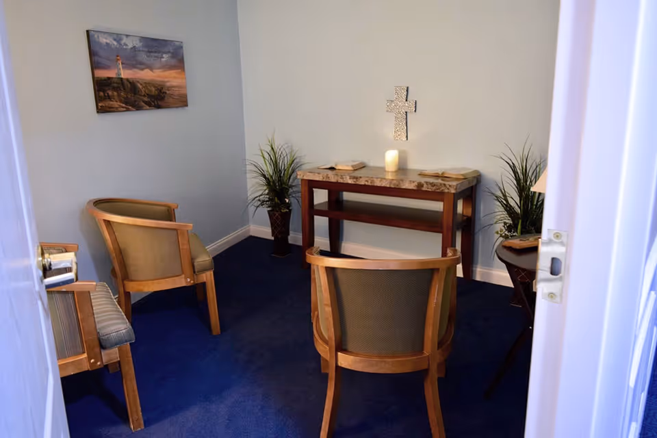 Small quiet room with blue carpet and light blue walls, containing three wooden chairs with cushions, a wooden table with a marble top holding an open book and a lit candle, two potted plants, a wall-mounted decorative cross, and a framed picture of a lighthouse on the wall.
